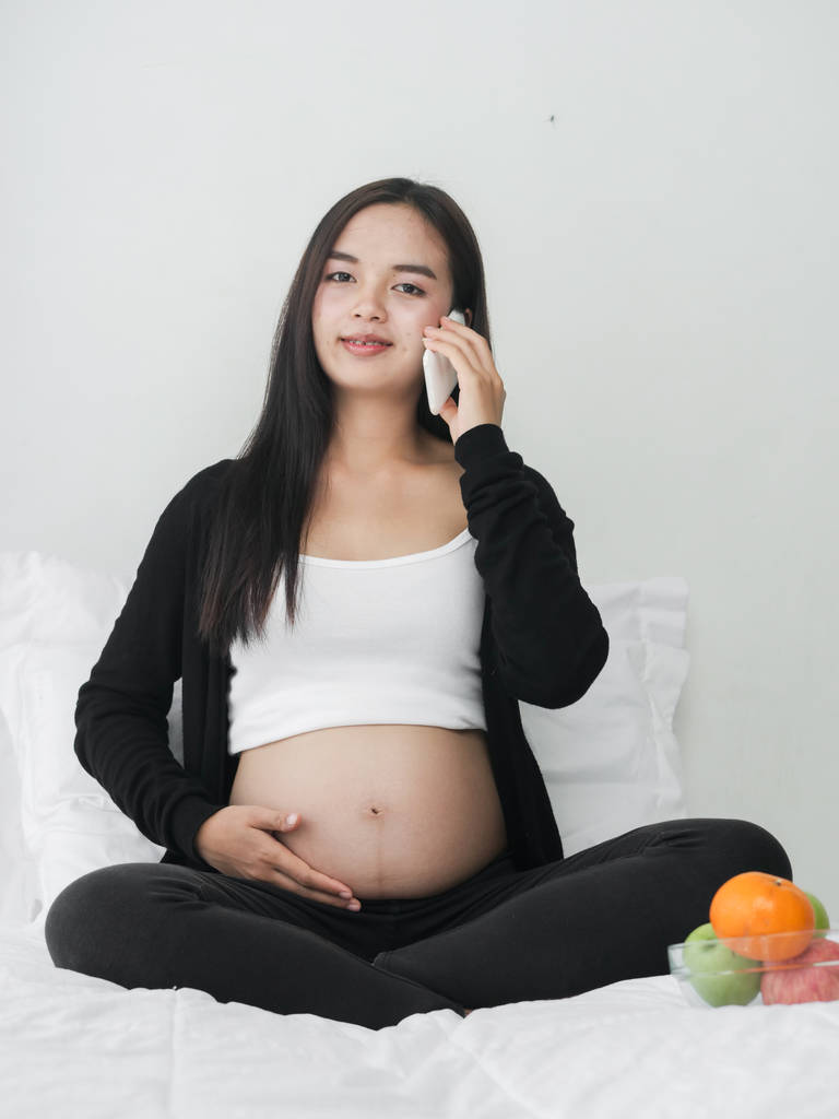 Szép ázsiai terhes nő használ szúró telefon ülve, ágyban, gyümölcsös tál, életmód fogalma. - Fotó, kép