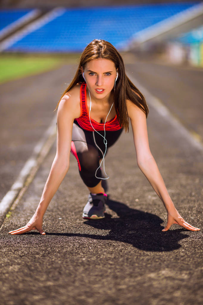 Портрет молодой спортсменки в спортивном платье, готовой к бегу и стоящей на старте на стадионной дорожке
 - Фото, изображение