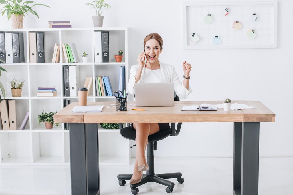 erfolgreiche Geschäftsfrau spricht auf Smartphone und triumphiert im modernen Büro mit Laptop - Foto, Bild