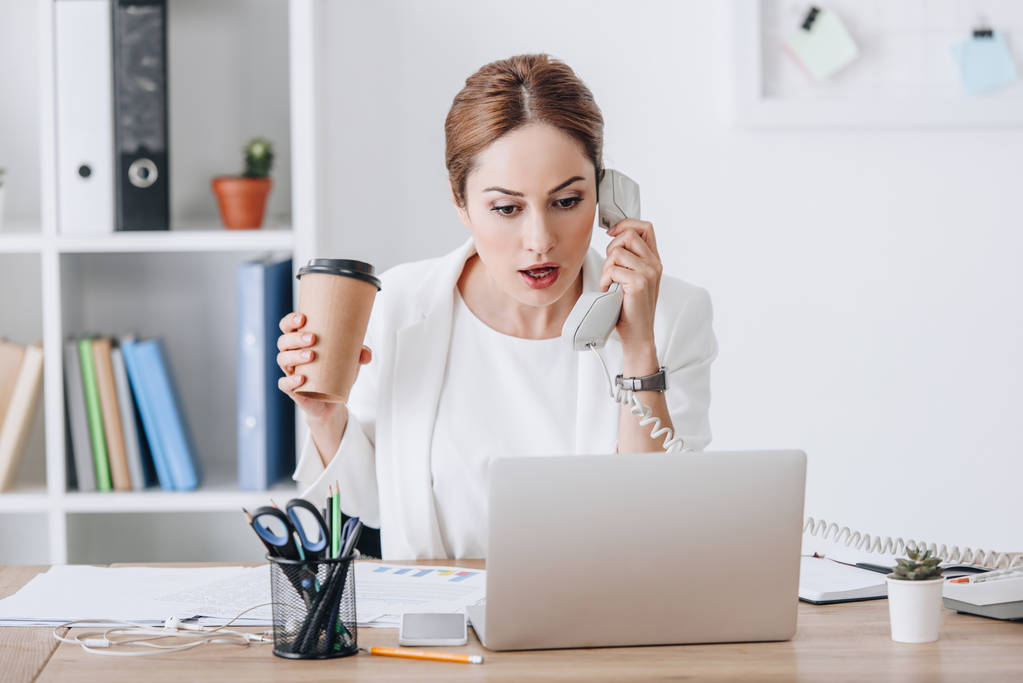красивая шокированная деловая женщина держит кофе, чтобы пойти во время разговора по телефону и работы с документами и ноутбуком на рабочем месте
 - Фото, изображение