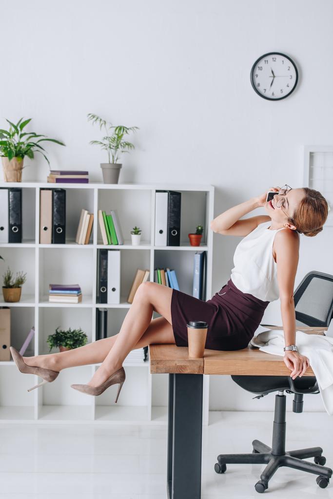 happy businesswoman talking on smartphone while sitting on table during coffee break in modern office - Photo, Image