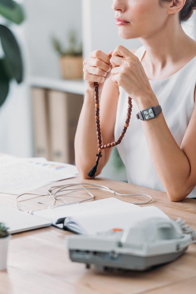 abgeschnittene Ansicht einer Geschäftsfrau, die im modernen Büro mit Rosenkranzperlen betet - Foto, Bild