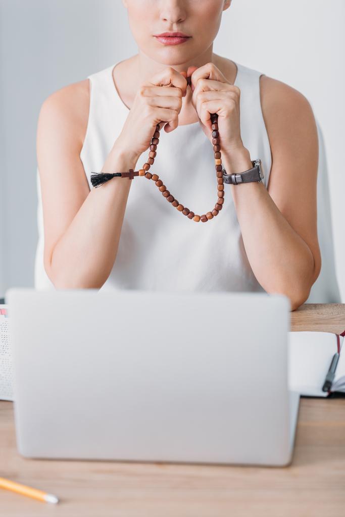 abgeschnittene Ansicht einer Geschäftsfrau, die mit Rosenkranzperlen betet und auf Laptop schaut - Foto, Bild