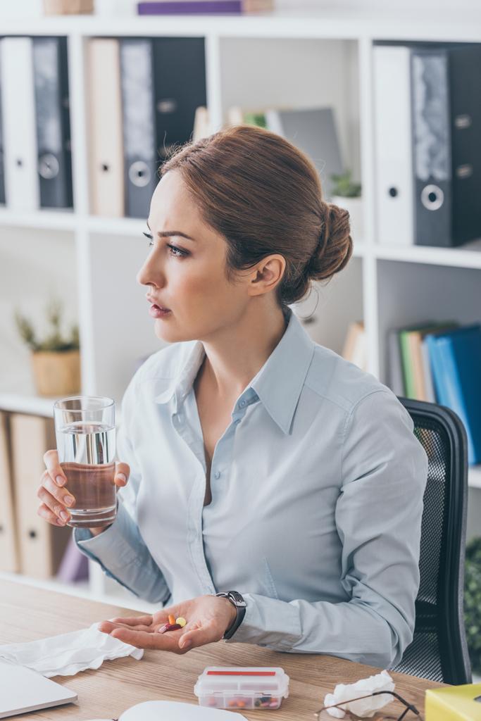 zamyšlený špatně podnikatelka sedí na pracovišti s prášky a sklenicí vody v kanceláři - Fotografie, Obrázek