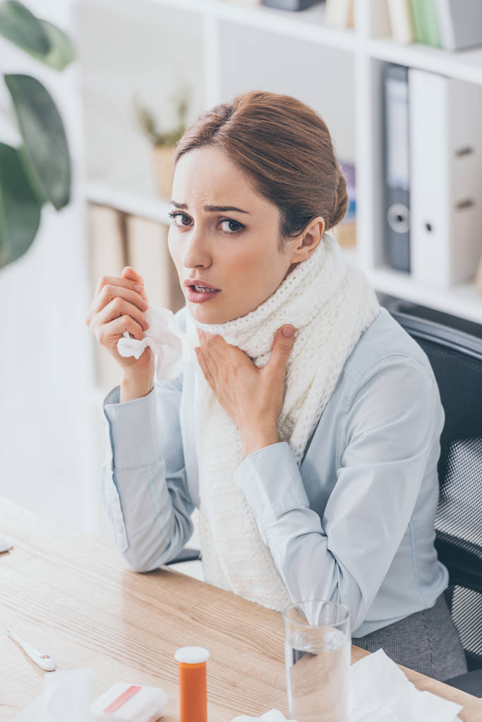 hoge hoekmening van zieke zakenvrouw in sjaal zit op de werkplek en camera kijken - Foto, afbeelding