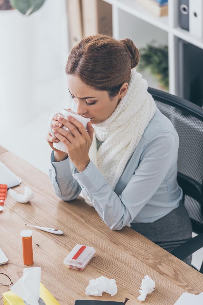 high angle view of sick businesswoman in scarf sitting at office and drinking tea - Photo, Image