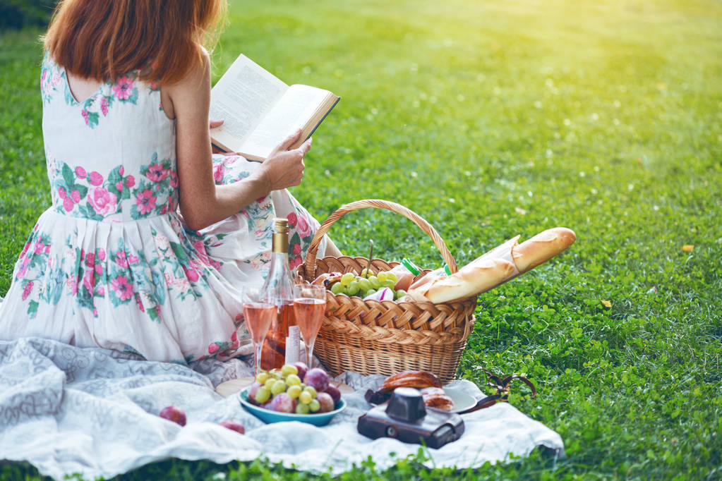 Verão - piquenique no prado. menina sentada lendo um livro e perto de uma cesta de piquenique e baguete, vinho, copos, uvas e rolo
 - Foto, Imagem