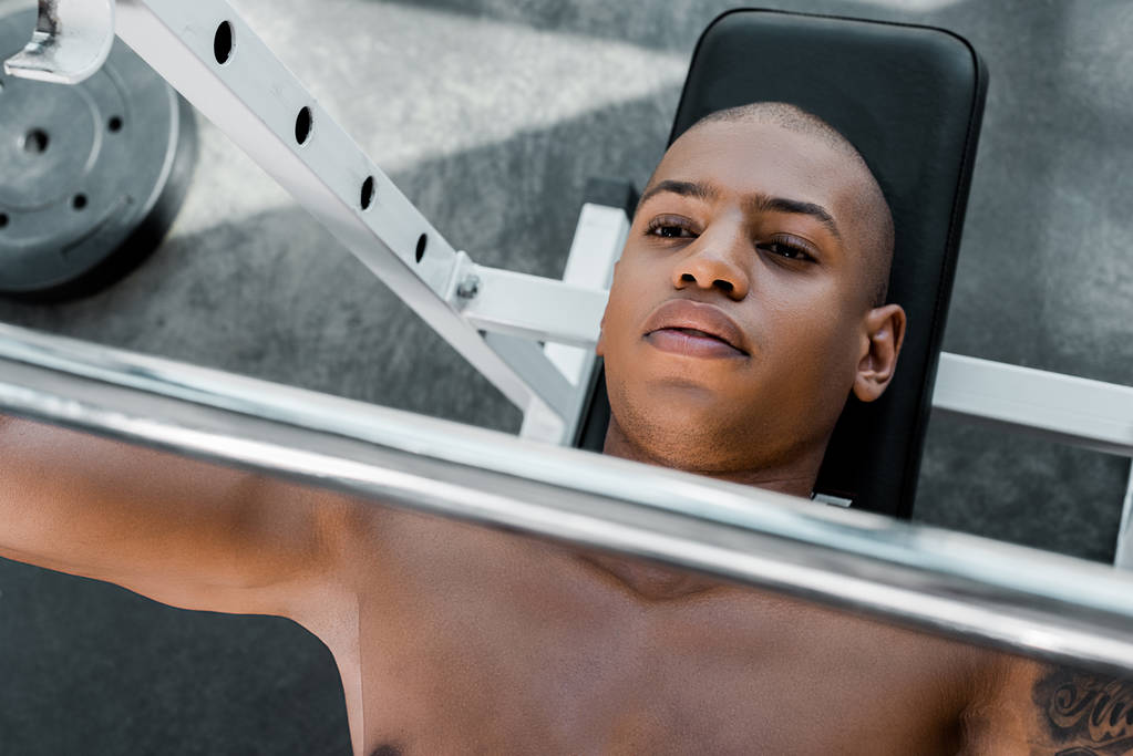 top view of young african american sportsman lying on bench and lifting barbell in gym - Photo, Image