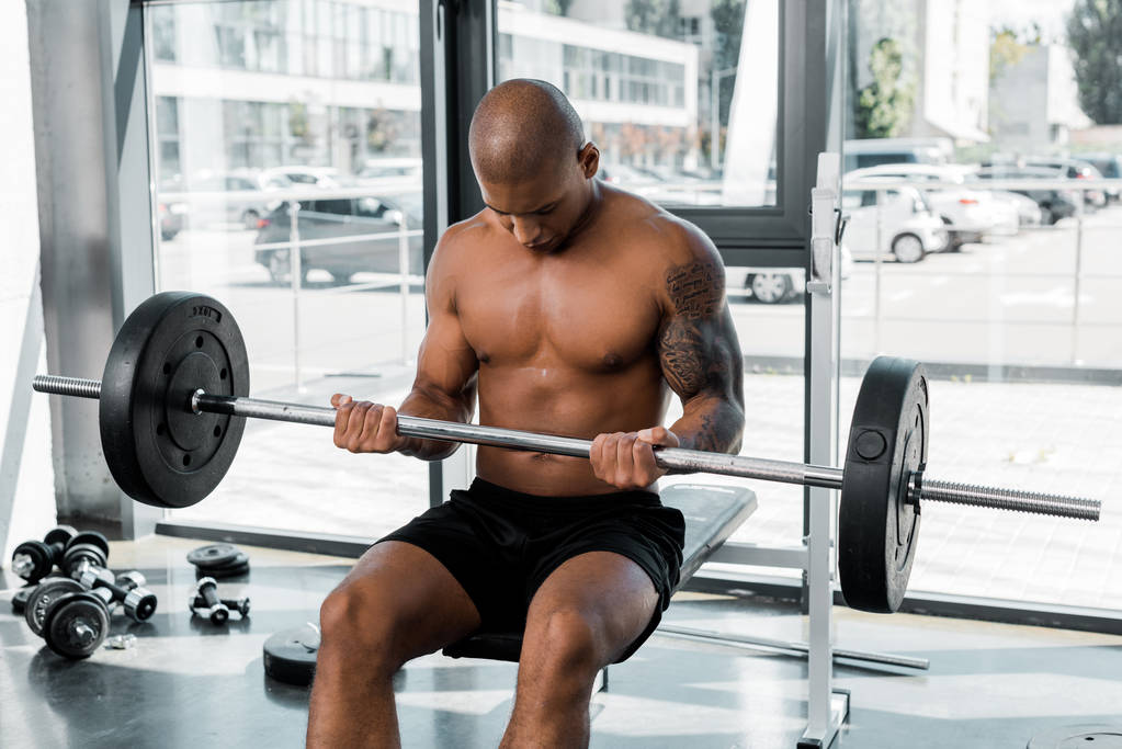 athletic bare-chested african american sportsman sitting on bench and lifting barbell in gym - Photo, Image