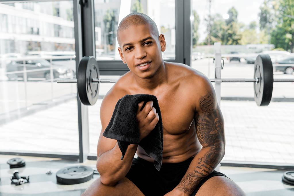 schöner muskulöser junger afrikanisch-amerikanischer Sportler mit Handtuch in der Hand und lächelnd in die Kamera im Fitnessstudio - Foto, Bild