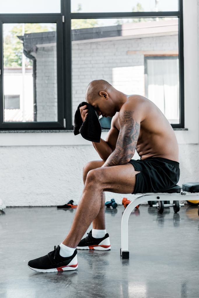 Widok z boku mięśni młody sportowiec ocierając czoło ręcznikiem siedząc po treningu w siłowni - Zdjęcie, obraz