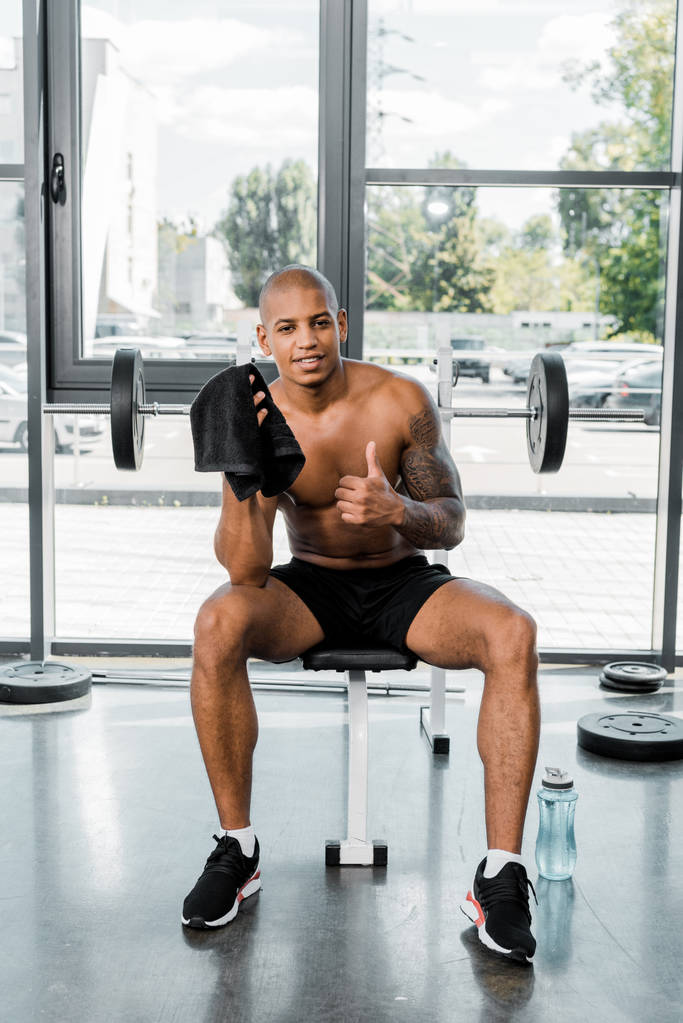 muskulöser junger afrikanisch-amerikanischer Sportler mit Handtuch, Daumen hoch und lächelnd in die Kamera im Fitnessstudio - Foto, Bild