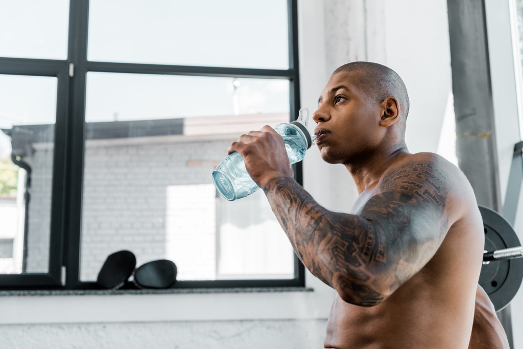 low angle view of muscular shirtless young tattooed sportsman drinking water in gym - Photo, Image