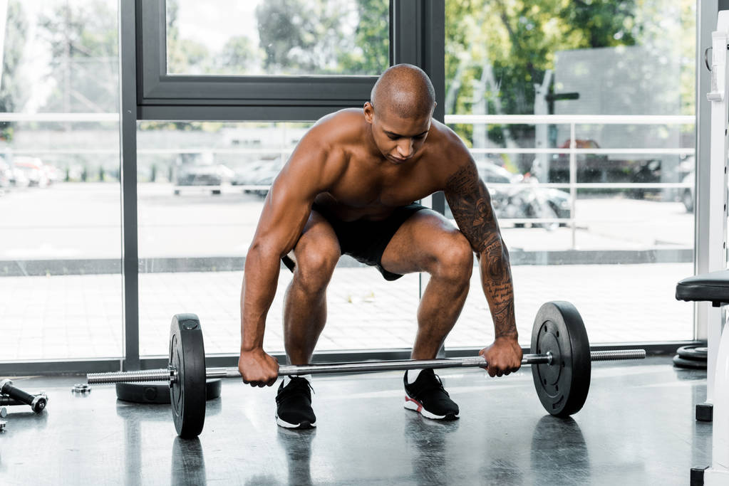 muskulöser junger afrikanisch-amerikanischer Mann mit nacktem Oberkörper beim Heben der Langhantel im Fitnessstudio - Foto, Bild