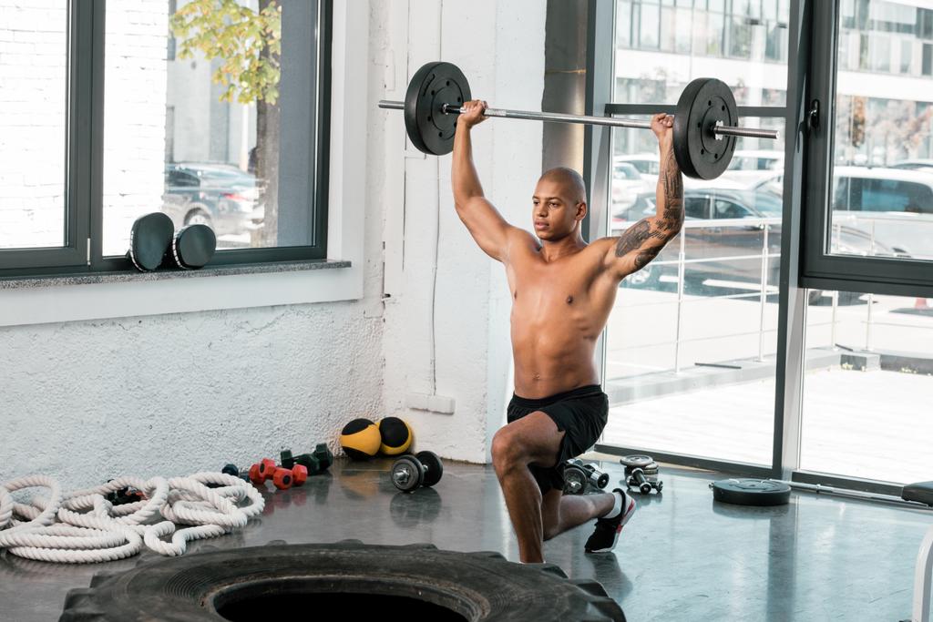 muskulöser junger Sportler mit nacktem Oberkörper, der die Langhantel hebt und im Fitnessstudio wegschaut  - Foto, Bild
