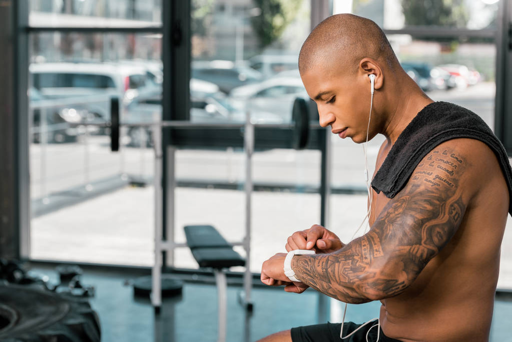 side view of young shirtless sportsman in earphones using smartwatch in gym  - Photo, Image