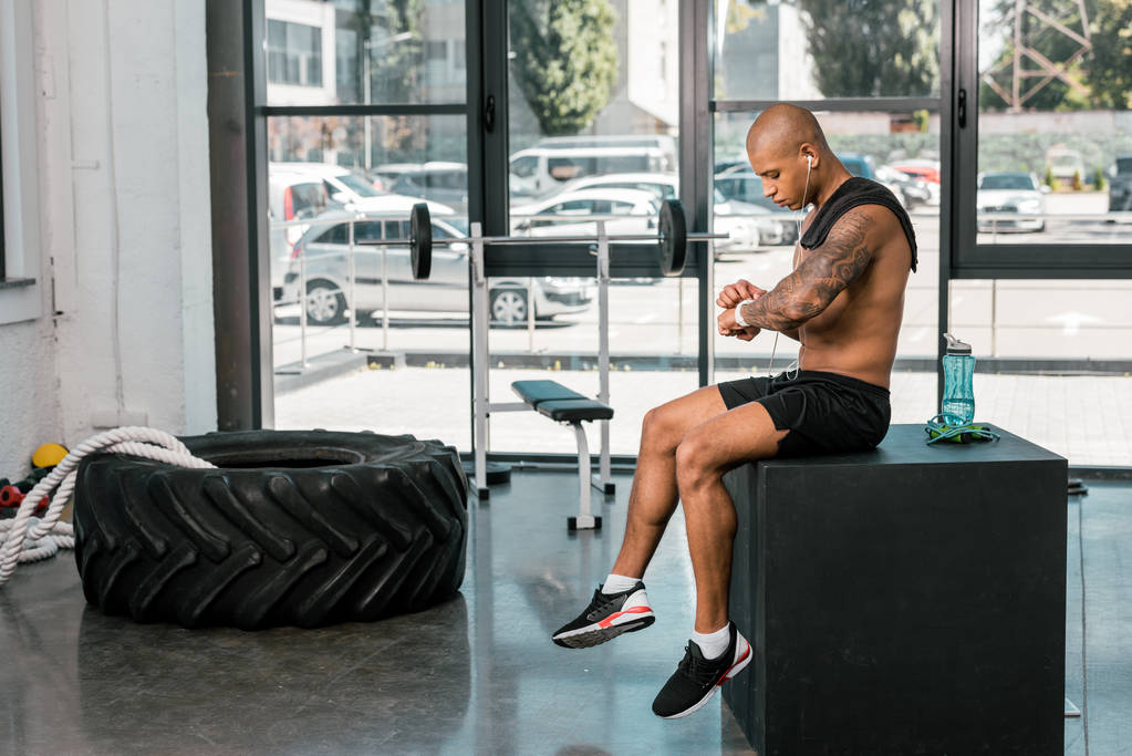 mięśni shirtless african american sportowca w słuchawki siedzi i za pomocą zegarka smartwatch w siłowni - Zdjęcie, obraz