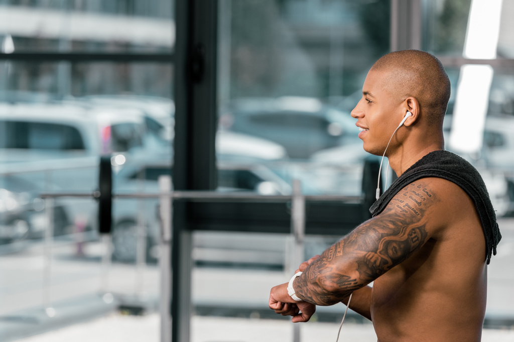 vue latérale du sportif torse nu souriant dans les écouteurs en utilisant smartwatch dans la salle de gym
  - Photo, image