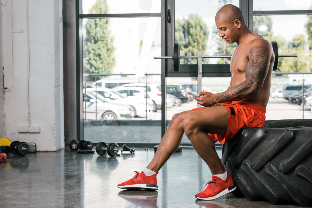 side view of happy african american man resting and using smartphone on tire at gym - Photo, Image