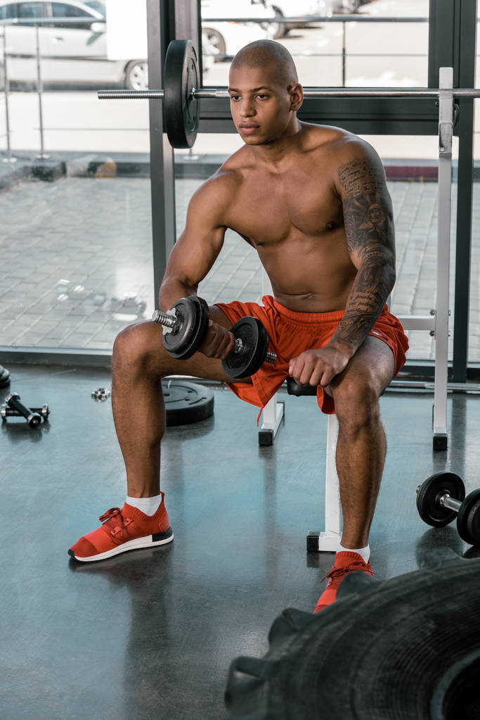 Американский спортсмен с татуировкой тренируется с гантелями в спортзале
 - Фото, изображение