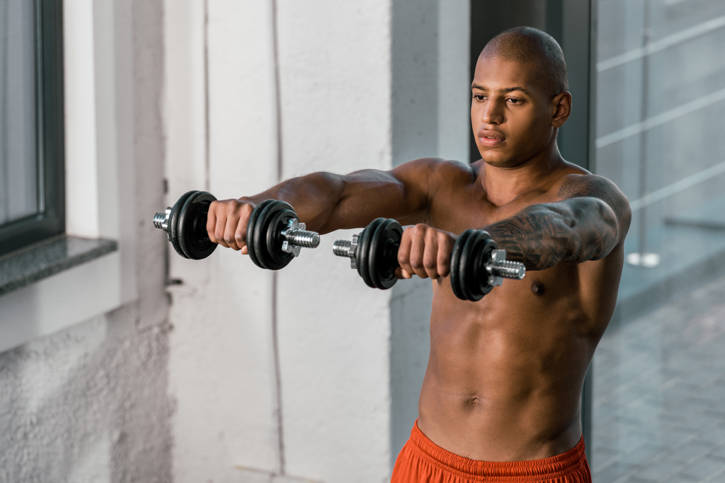 young muscular shirtless african american sportsman exercising with dumbbells at gym - Photo, Image