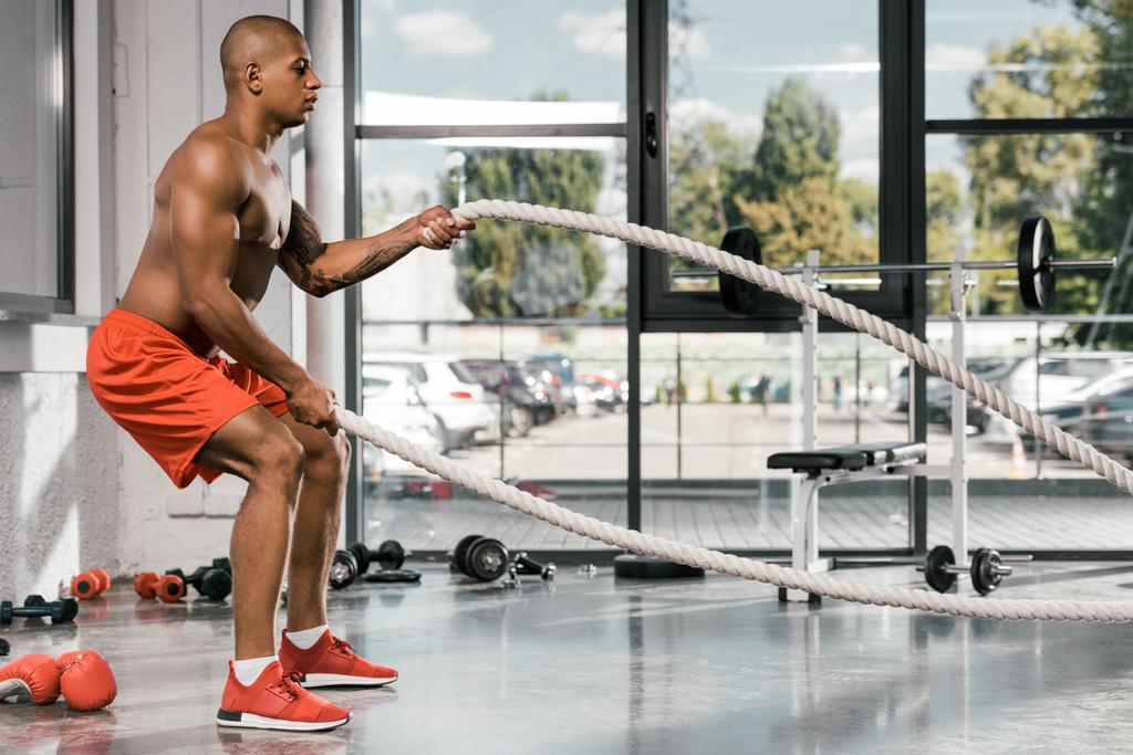 Seitenansicht eines afrikanisch-amerikanischen Athleten, der im Fitnessstudio mit Kampfseilen trainiert - Foto, Bild