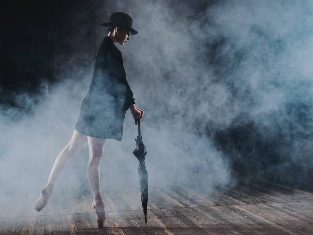 Ballerina in black long jacket and sexy bodysuit dancing on stage with magic light and smoke. Young attractive dancer in ballet shoes pointe with umbrella - Photo, Image