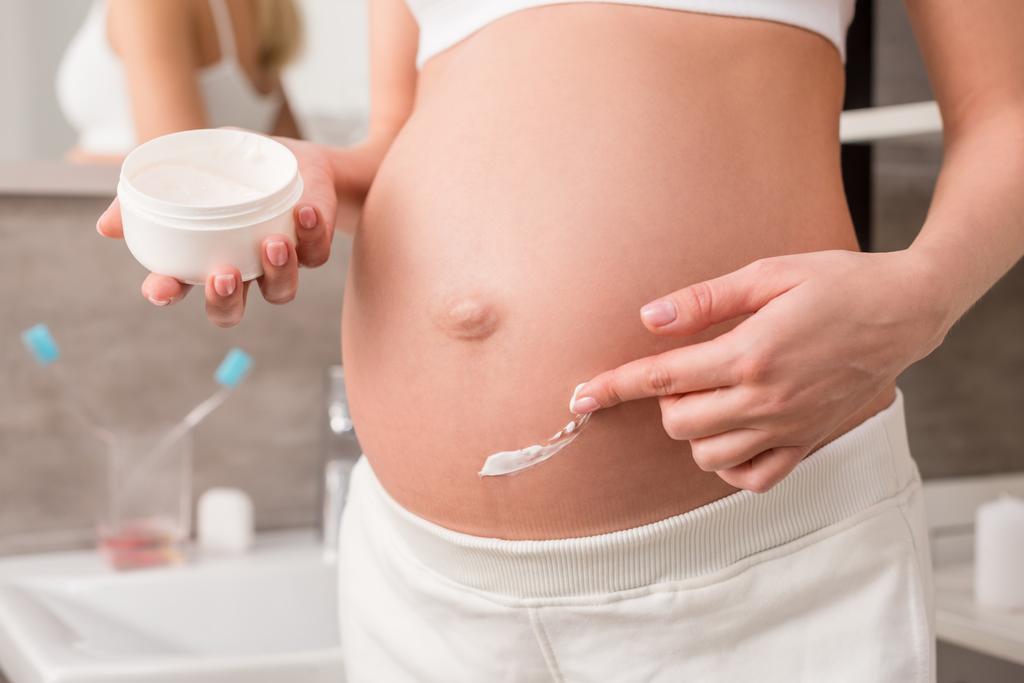 обрізаний знімок вагітної жінки, що наносить крем на животик, щоб уникнути розтяжок у ванній
 - Фото, зображення