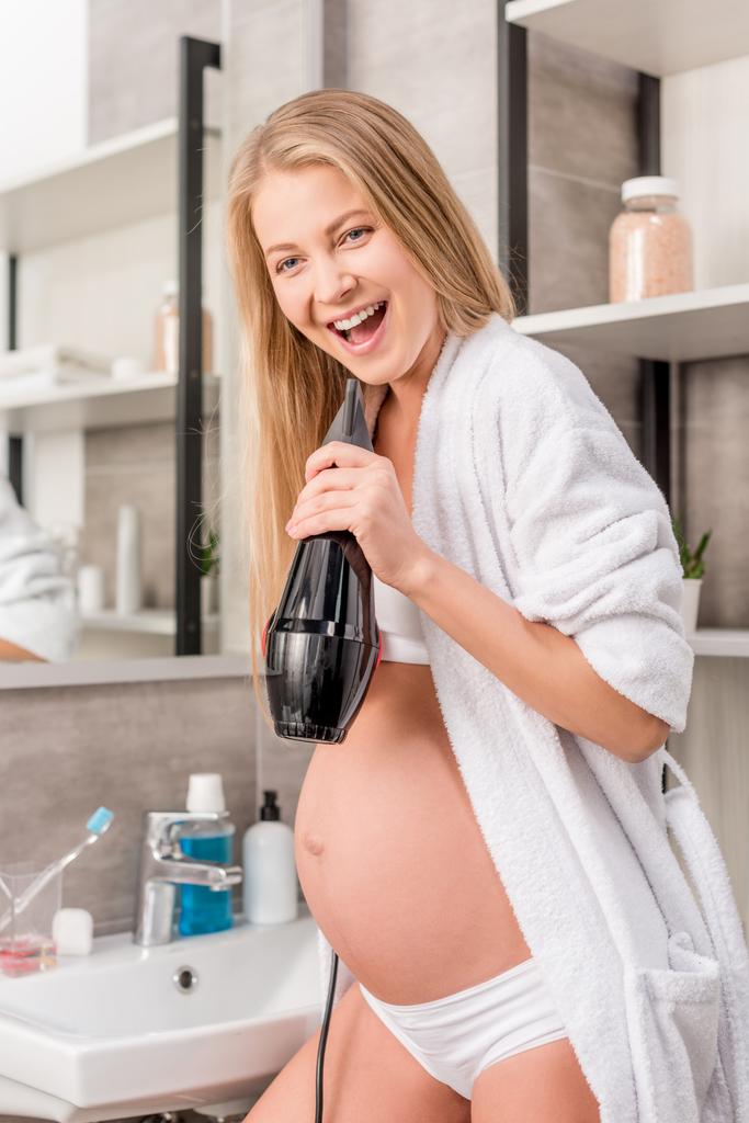 Gelukkig zwangere vrouw zingen op een haardroger in de badkamer - Foto, afbeelding