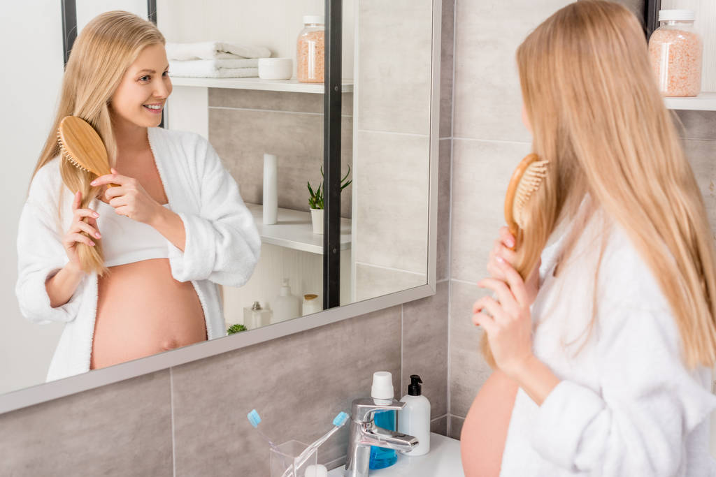 heureuse femme enceinte en peignoir brossant ses cheveux et regardant miroir dans la salle de bain
 - Photo, image