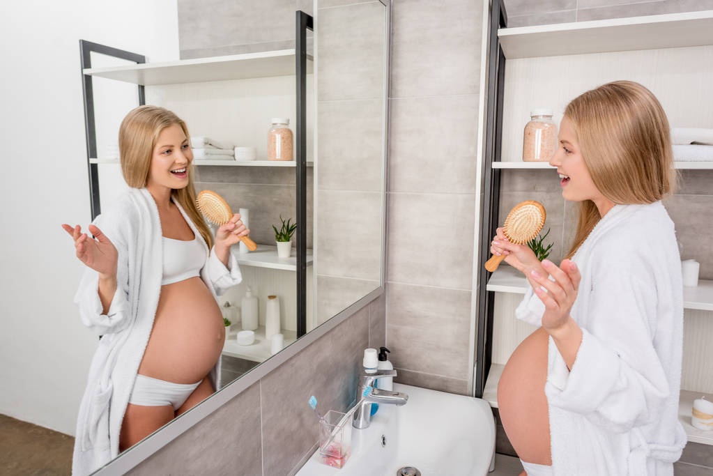 Szczęśliwa Kobieta w ciąży w bieliźnie, śpiewając w szczotkę i patrząc w lustro w łazience - Zdjęcie, obraz