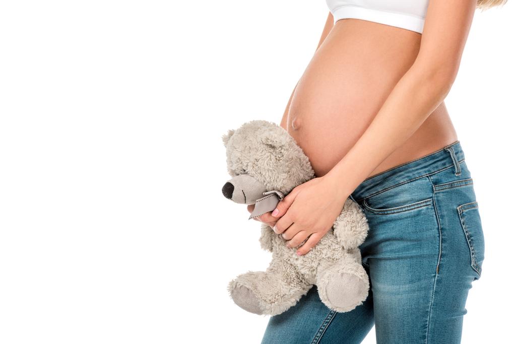 obcięty widok kobiety w ciąży, trzymając pluszowego misia w pobliżu jej brzuch na białym tle - Zdjęcie, obraz