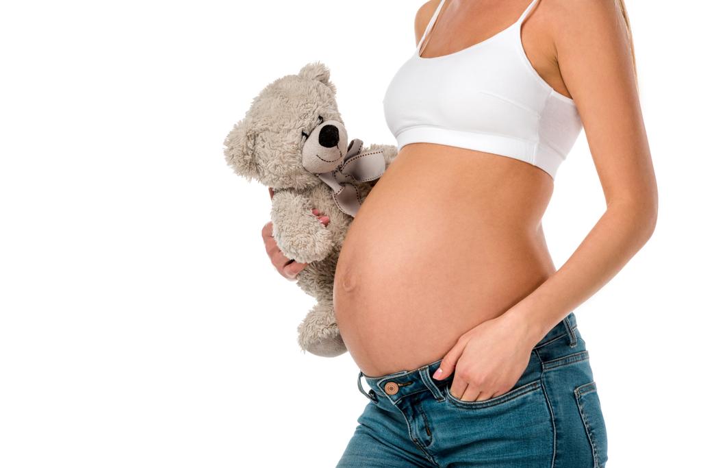abgeschnittene Ansicht von schwangeren Mädchen in weißen BH hält Teddybär isoliert auf weiß - Foto, Bild