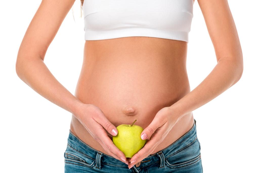 abgeschnittene Ansicht einer schwangeren Frau, die einen gesunden Apfel in der Nähe ihres Bauches hält, isoliert auf weiß - Foto, Bild