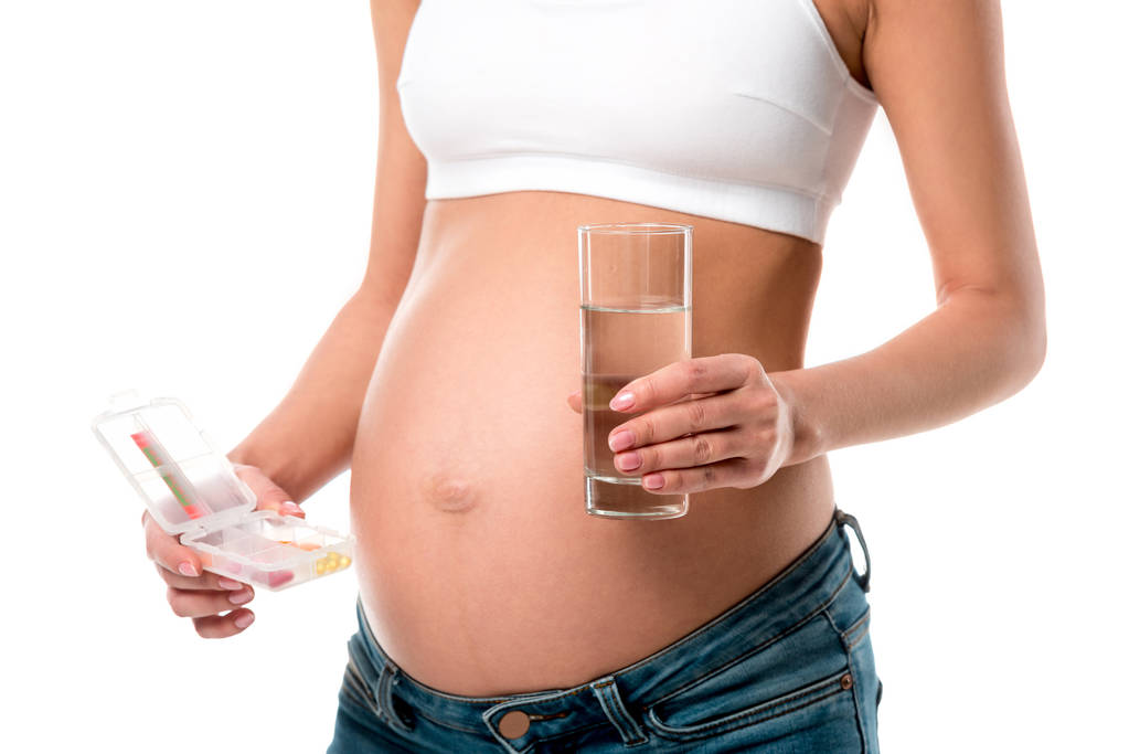 περικοπεί άποψη της εγκύου γυναίκας κρατώντας κουτί χάπι και ποτήρι νερό που απομονώνονται σε λευκό - Φωτογραφία, εικόνα