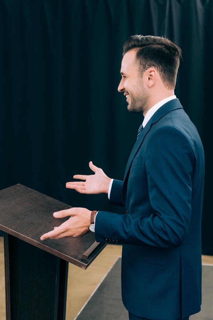 vista lateral del apuesto conferenciante de pie y haciendo gestos en el podio tribuno durante el seminario en la sala de conferencias
 - Foto, Imagen