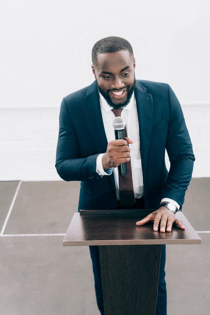 korkea kulma näkymä hymyilevä afrikkalainen amerikkalainen puhuja puhuu mikrofoniin seminaarin aikana konferenssisalissa
 - Valokuva, kuva