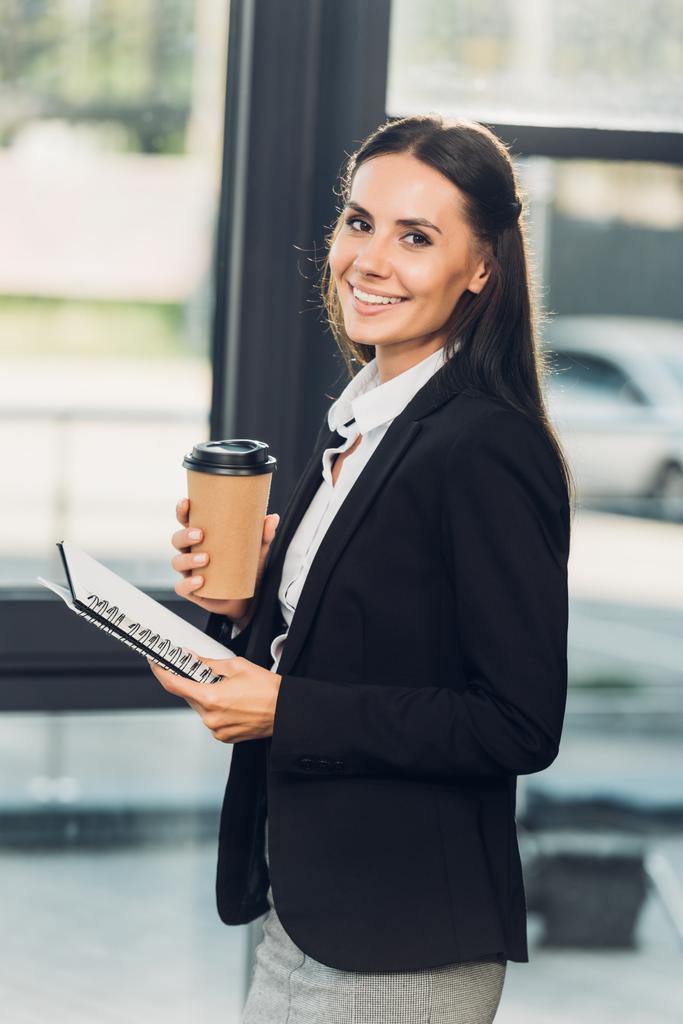 vista lateral da jovem mulher de negócios sorridente com café para ir e notebook na sala de conferências
 - Foto, Imagem