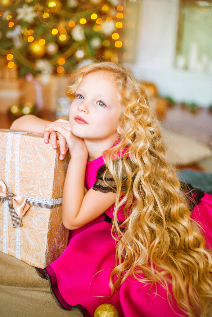 Menina bonito com cabelo louro encaracolado longo em casa perto de uma árvore de Natal com presentes e guirlandas e uma lareira decorada
  - Foto, Imagem