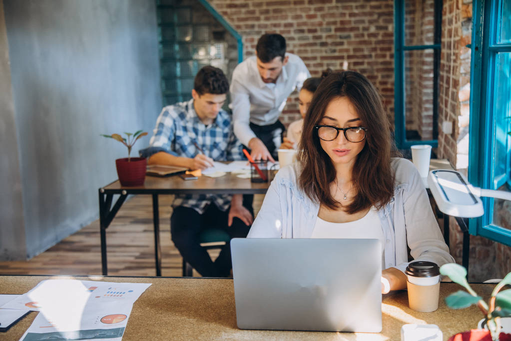Молодая бородатая деловая женщина в джинсовой рубашке сидит в офисе за столом и использует ноутбук с графиками, графиками и диаграммами на экране. Человек работает. startu
 - Фото, изображение