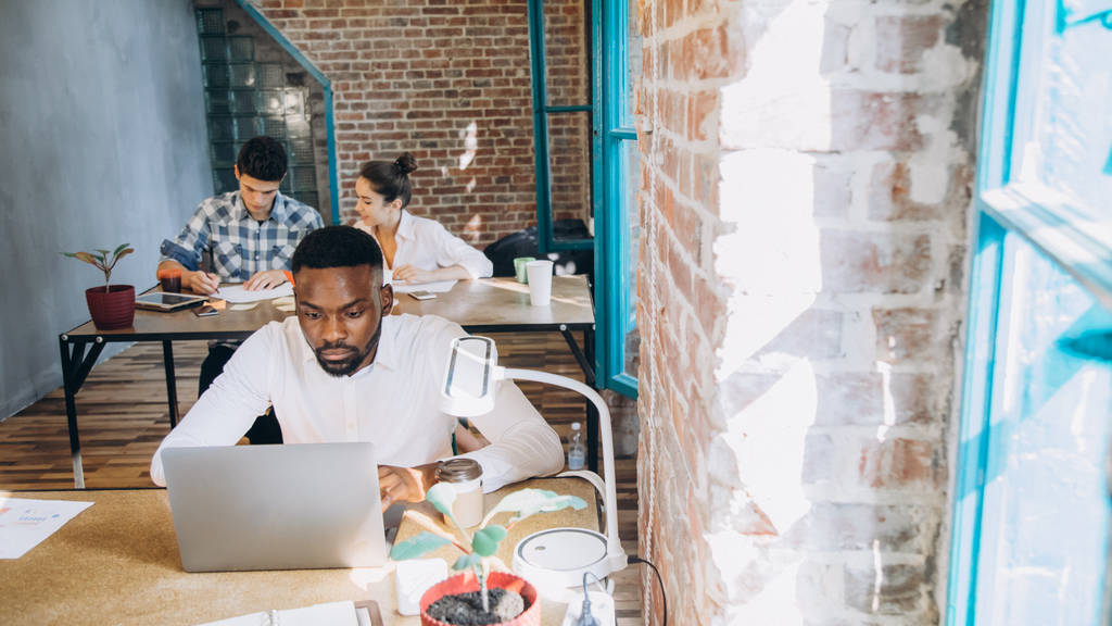 uomo d'affari afroamericano premuroso seduto al tavolo e colleghi che lavorano dietro in ufficio moderno. startup
 - Foto, immagini