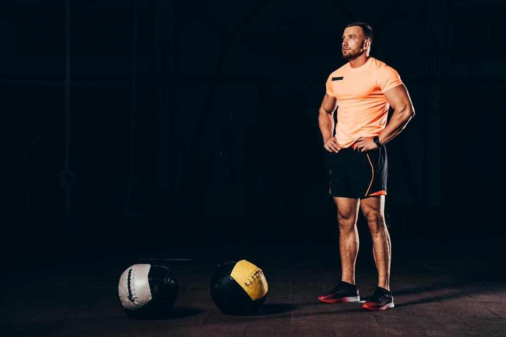 schöner athletischer Bodybuilder steht in dunkler Turnhalle mit Medizinbällen am Boden - Foto, Bild
