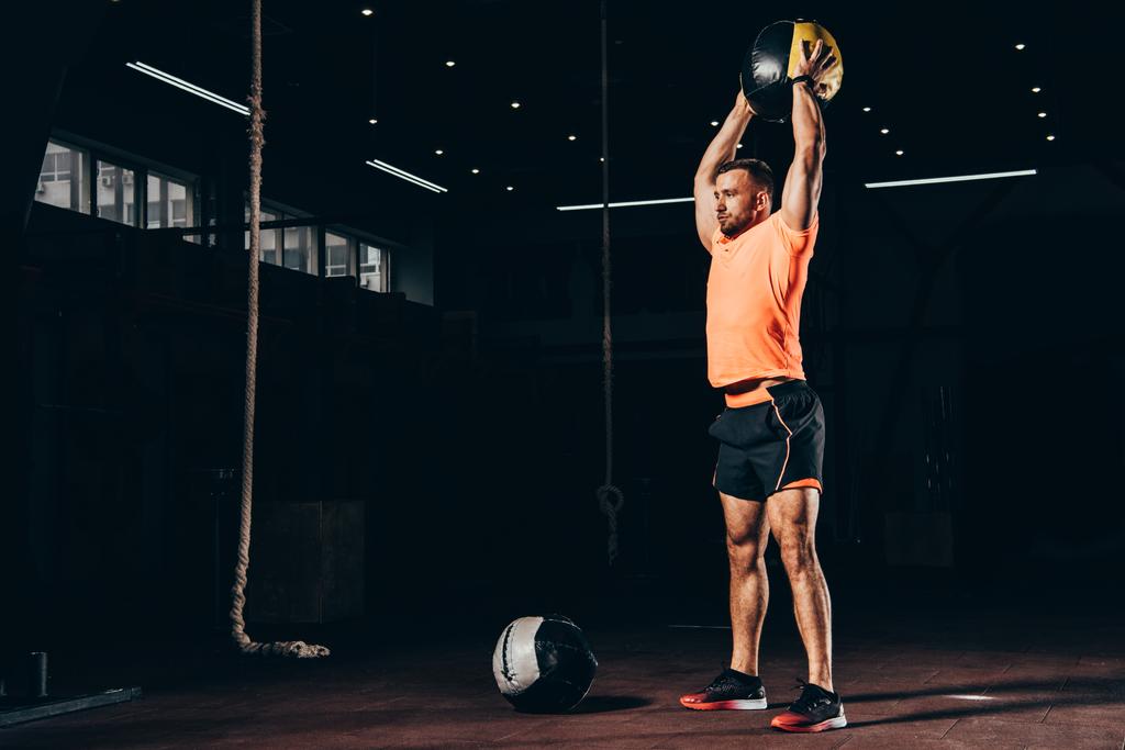 handsome fit sportsman standing with medicine ball overhead in dark gym - Photo, Image