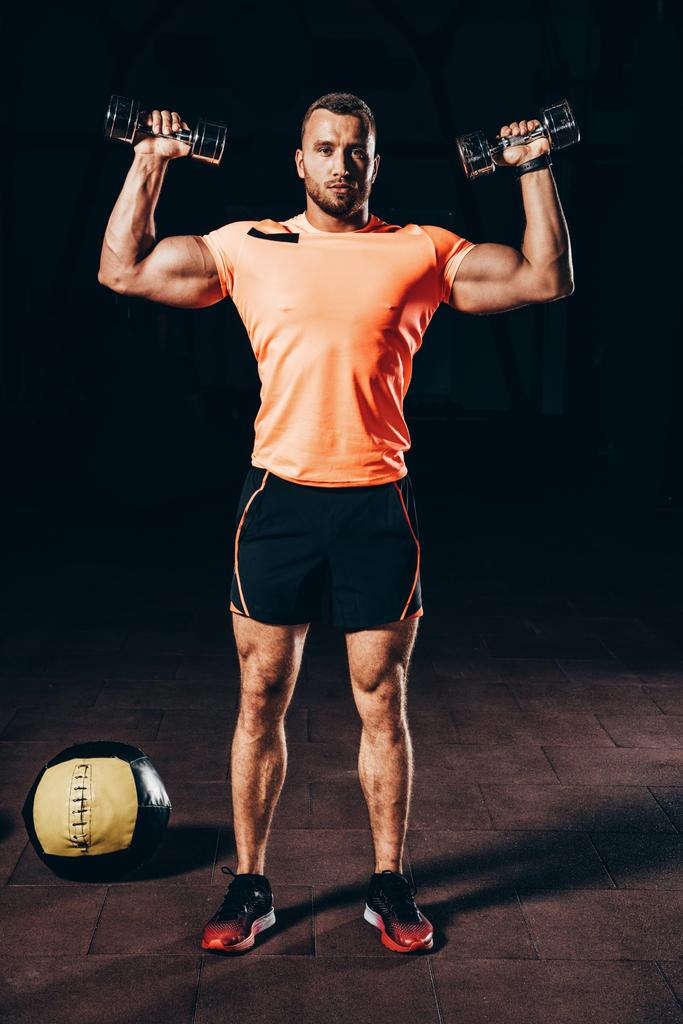 όμορφος bodybuilder που εργάζονται έξω με αλτήρες σε σκούρο γυμναστήριο - Φωτογραφία, εικόνα