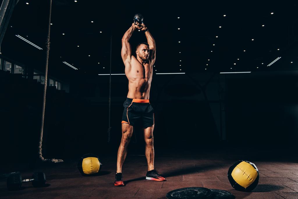 apuesto deportista en forma sosteniendo kettlebell sobre la cabeza mientras se entrena en el gimnasio oscuro
 - Foto, Imagen