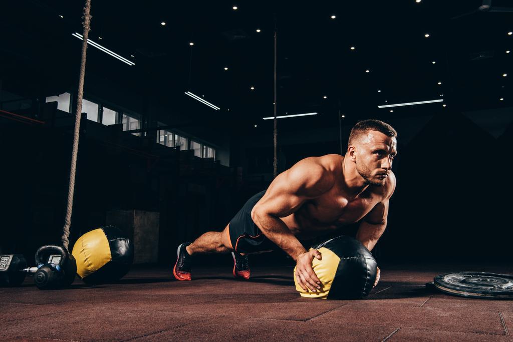 όμορφος αθλητικός αθλητής κάνει push ups στην ιατρική μπάλα στο σκοτεινό γυμναστήριο - Φωτογραφία, εικόνα