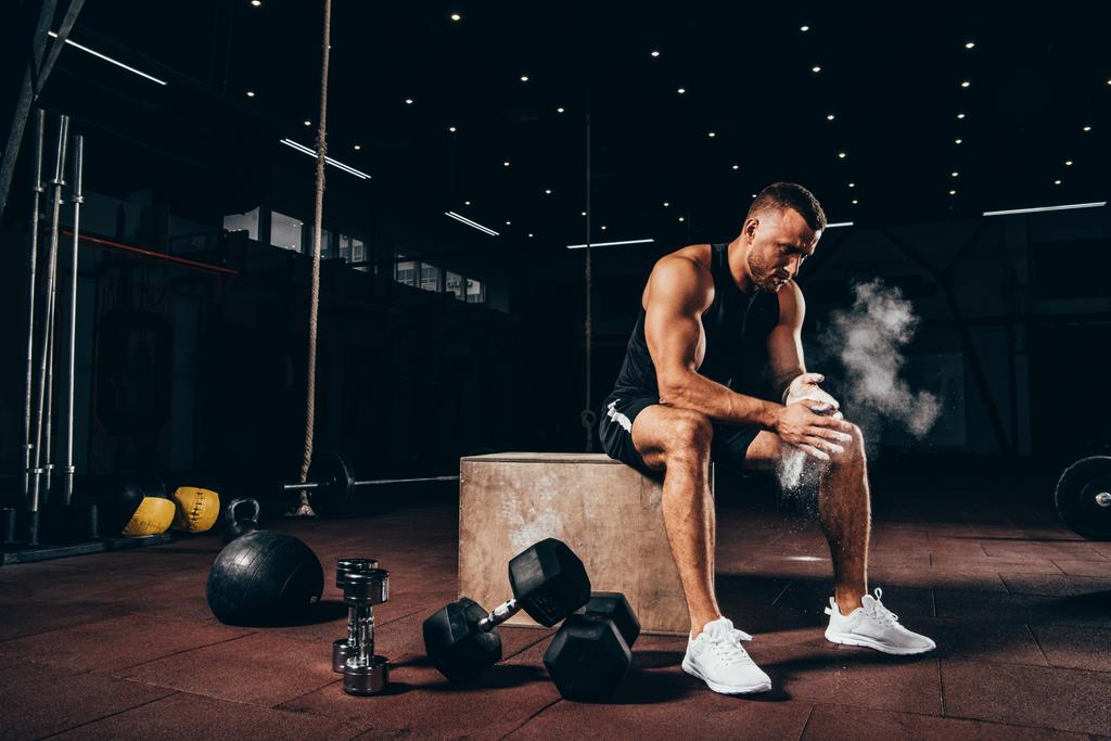 handsome athletic man sitting on cube with gym equipment around and clapping hands with talc before workout - Photo, Image