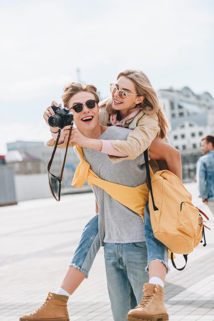 happy boyfriend giving piggyback to girlfriend with photo camera in city - Photo, Image