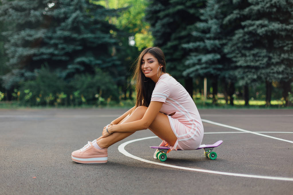 Πορτρέτο ενός χαμογελαστού γοητευτικό μελαχρινή γυναίκα κάθεται στο skateboard της σε ένα γήπεδο μπάσκετ. Ευτυχισμένη γυναίκα με μοντέρνα εμφάνιση λαμβάνοντας διάλειμμα κατά τη διάρκεια του ηλιοβασιλέματος. - Φωτογραφία, εικόνα