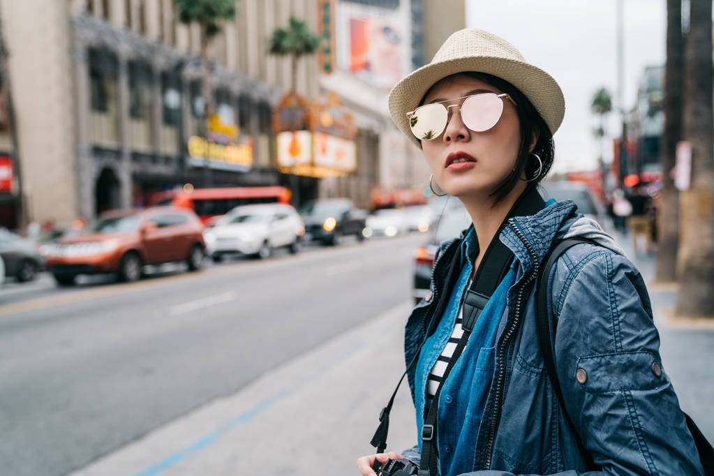 Κομψό γυναικείο ταξιδιώτη στέκεται στο δρόμο και να περιμένει ταξί να πάει πίσω στο ξενοδοχείο. γυναίκα σε ψάθινο καπέλο και γυαλιά ηλίου που βλέπουν στο δρόμο. δροσερό καιρό στο La. - Φωτογραφία, εικόνα